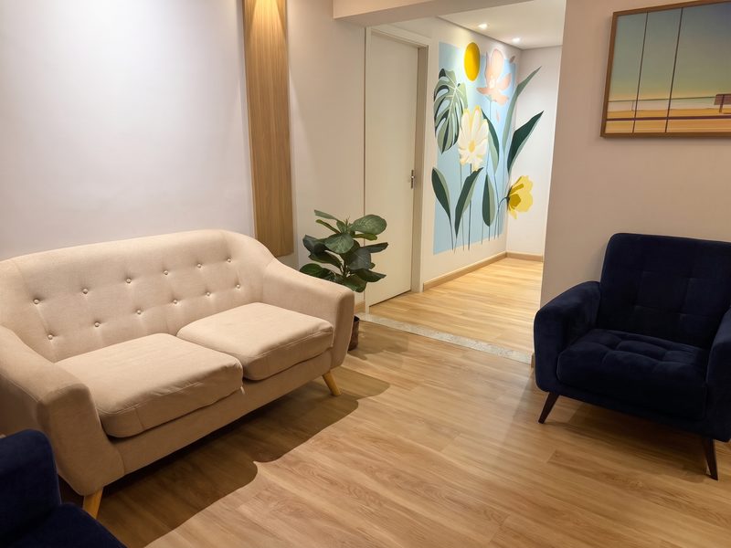 Sala de espera do consultório, com sofá bege, poltronas azuis e mural de flores